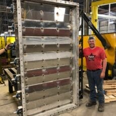 A Kelair shop worker smiles while standing next to a massive louver damper that is twice his size.
